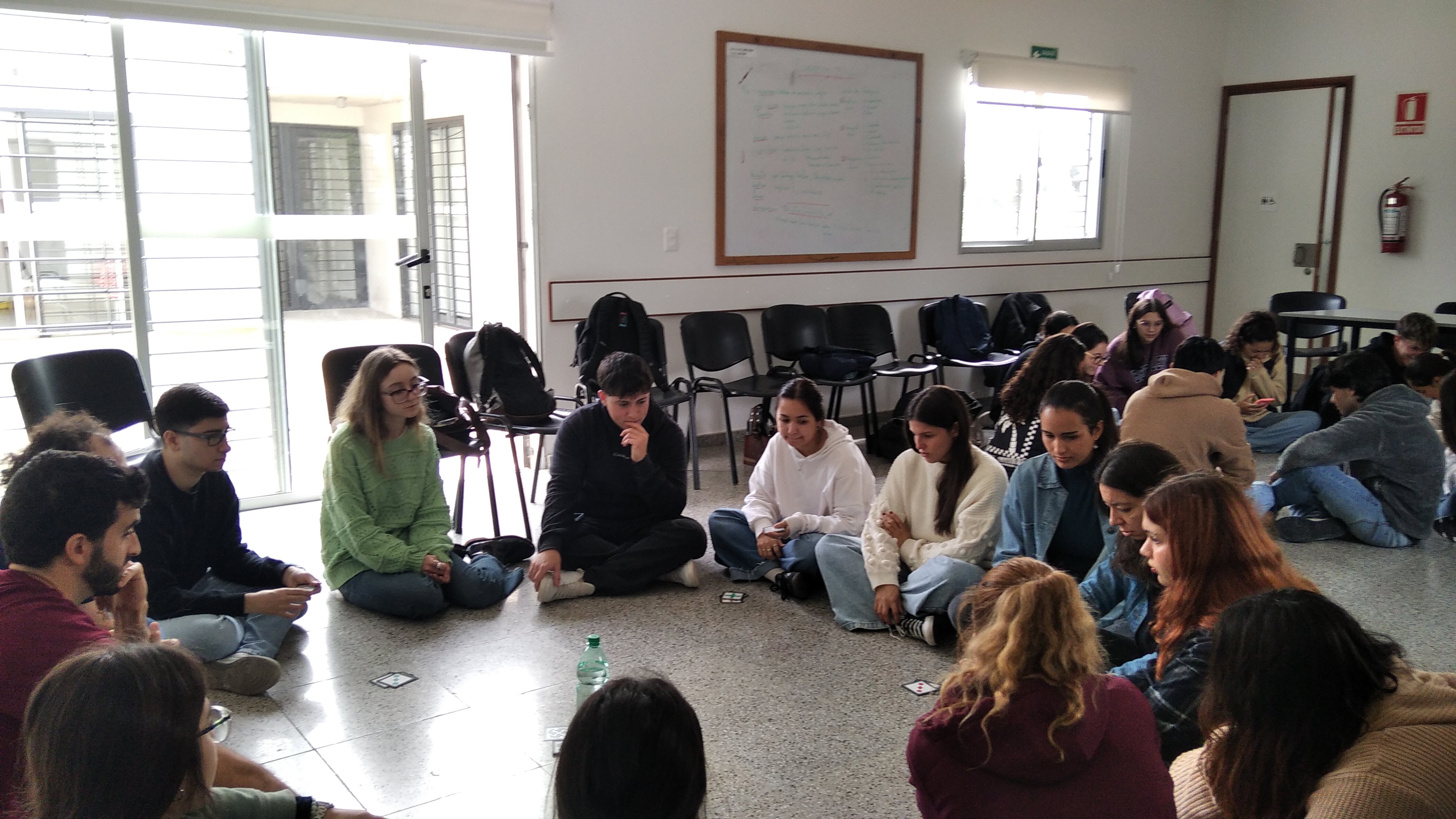 En la imagen se puede observar un salón de clase donde hay dos grupos de jóvenes que estan en ronda sentados con cartas.